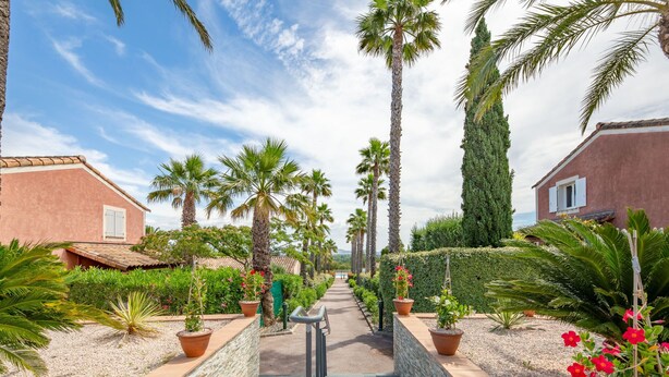 A pathway flanked by palm trees and potted plants, leading to a building with a red facade and a tiled roof. - Le Green Village by Interhome (Roquebrune sur Argens)