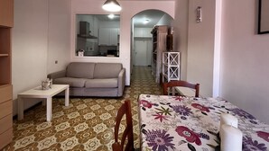 Un espai vital compacte amb un sofà, una taula petita, una taula de menjador amb un mantell de flors, una làmpada penjant i una cuina al fons.