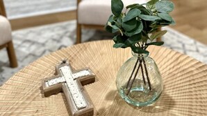 A living room with a white fireplace, a beige armchair, a round coffee table with a glass vase and a wooden cross, and a kitchen in the background. - Fort Smith / Furnished 4 BR / Nurses, Ebbing, TDY (Fort Smith)