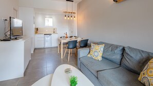 Una sala de estar moderna con un sofá gris, una mesa de centro y un televisor sobre un mueble. Hay una zona de comedor con una mesa y sillas, y una cocina al fondo.