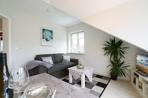 Een moderne woonkamer met een grijze bank, een klein tafeltje, een potplant en een televisie.