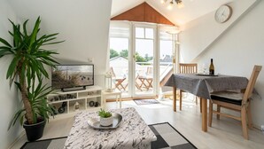 Una sala de estar acogedora con televisor, una mesa de comedor con sillas, una planta en maceta y vistas al exterior a través de un ventanal.