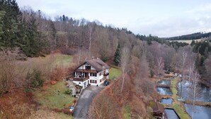Une maison à deux étages dotée d’un balcon, entourée d’une zone boisée et d’un petit étang.