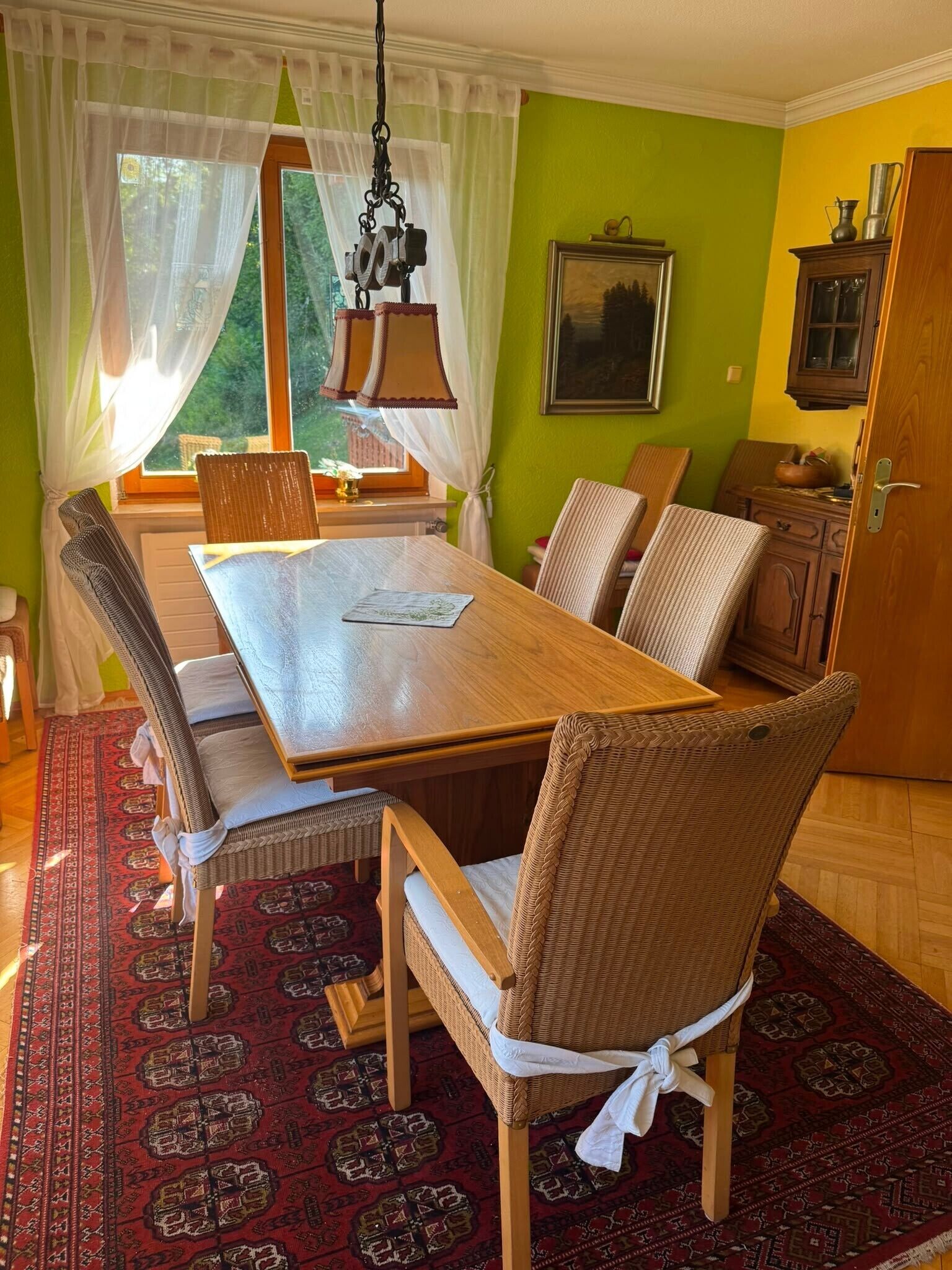 En spisestue med et træbord, fletstole, et mønstret tæppe og en hængende lampe.