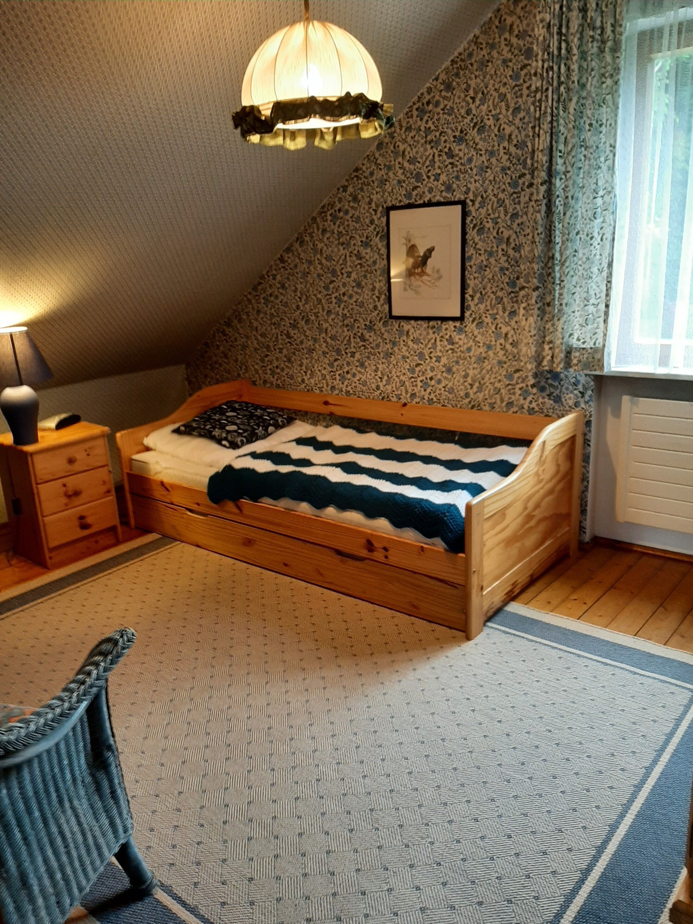 Ein Schlafzimmer mit einem hölzernen Bett, einem Nachttisch, einer Lampe, einem gerahmten Bild an der Wand und einem Fenster mit Vorhängen.