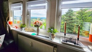 A room with three windows, a radiator, a small table with a vase of flowers, and a decorative ship model. - Apartment Waldblick with Large Balcony, Ground-Level Access, and Unique Forest View (Bestwig)