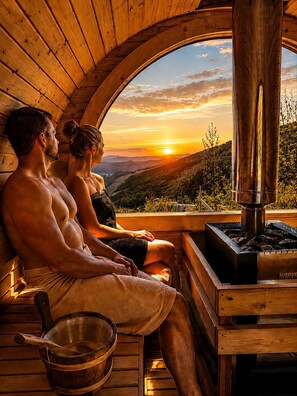 A couple relaxing in a wooden cabin with a sauna, a view of the sunset, and a wooden table with a bowl and spoon.