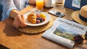 Una persona sentada a una mesa con un plato de comida, un vaso de jugo de naranja, un periódico y una ventana con vista a un paisaje.