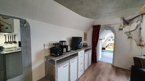 Una cocina compacta con cafetera, tostadora y televisor sobre un mostrador, un lavamanos y armarios. Un arco de entrada da a un dormitorio.