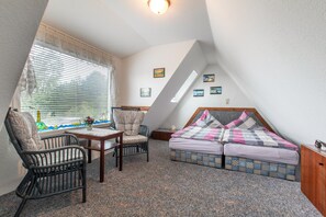 Un dormitorio acogedor en el ático, con techo inclinado, una cama con colcha estampada, dos sillas, una mesita y una ventana con persianas.