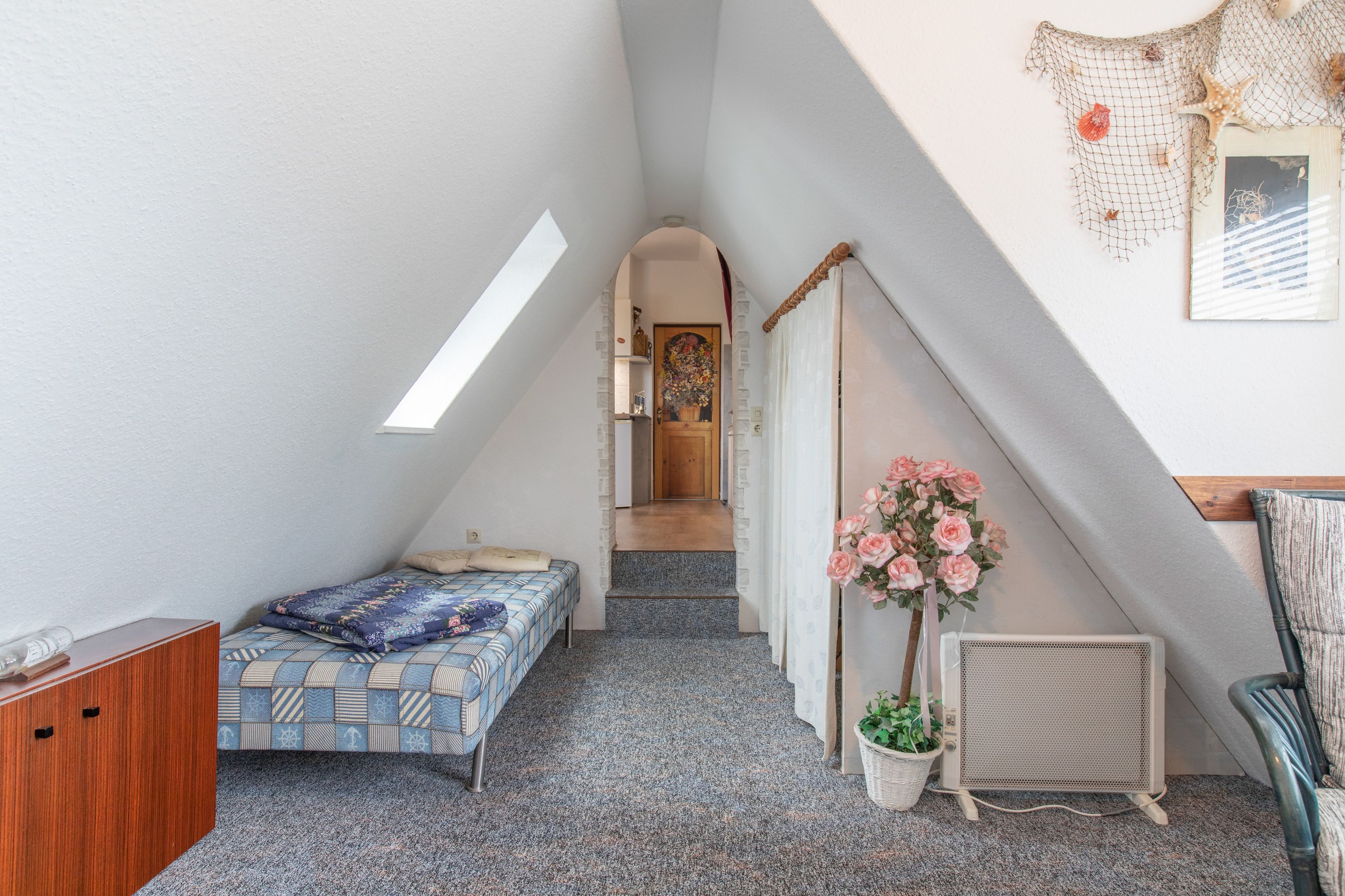 Un dormitorio compacto con techo inclinado, una cama con ropa de cama estampada, una planta en maceta, un radiador y un pequeño armario de madera.