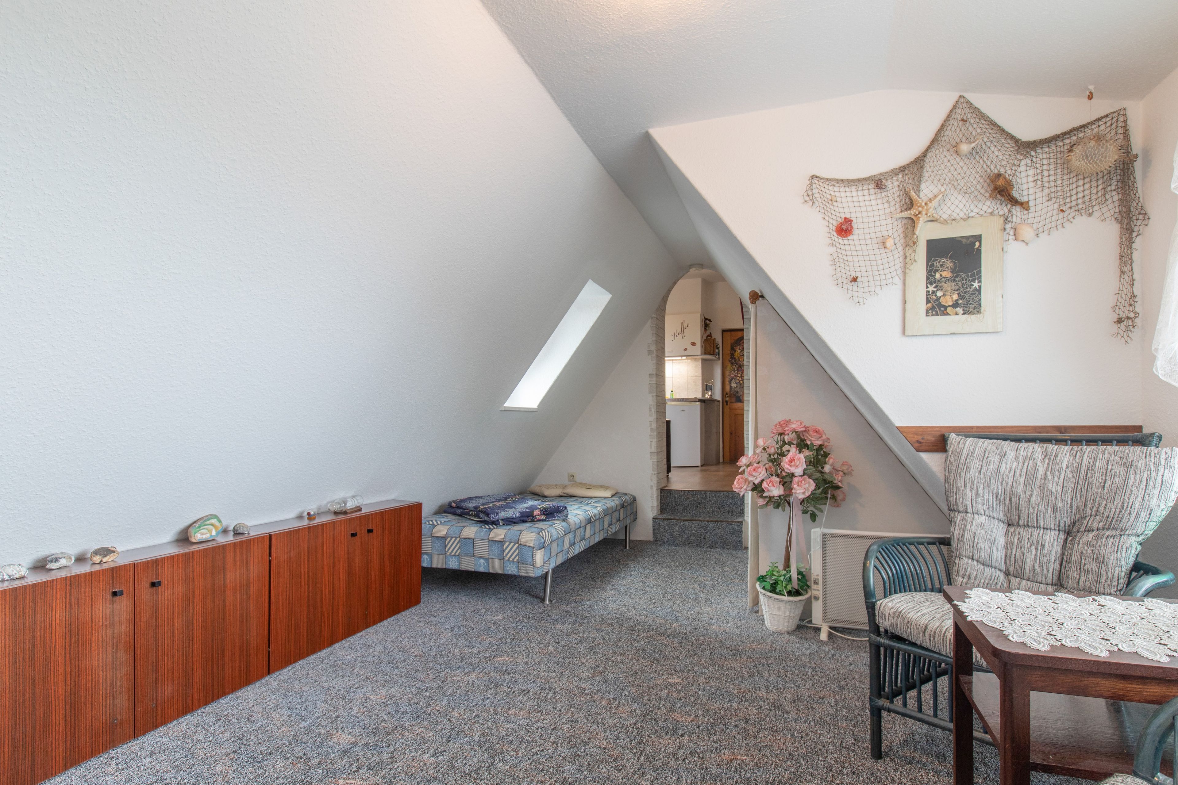 Une chambre mansardée chaleureuse, avec un plafond en pente, une armoire en bois, un lit recouvert d’une couette à motifs, une chaise, une petite table avec un chemin de table en dentelle et une étagère murale agrémentée d’objets décoratifs.