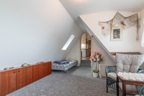 A cozy attic bedroom with a sloped ceiling, a wooden wardrobe, a bed with a patterned duvet, a chair, a small table with a lace tablecloth, and a wall-mounted shelf with decorative items.