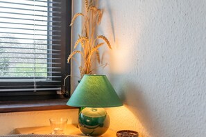 Une lampe avec un abat-jour vert, un vase contenant des plantes séchées et une fenêtre avec des stores.