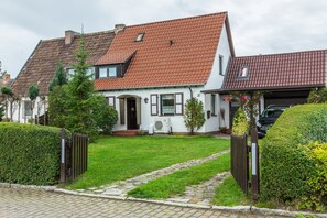Une maison à deux étages avec un toit recouvert de tuiles rouges, une cour avant avec un chemin pavé et une voiture noire garée dans l’allée.