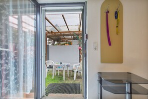 Puerta abierta que da a un patio con mesa y sillas, un pequeño jardín y un estante en la pared con objetos.