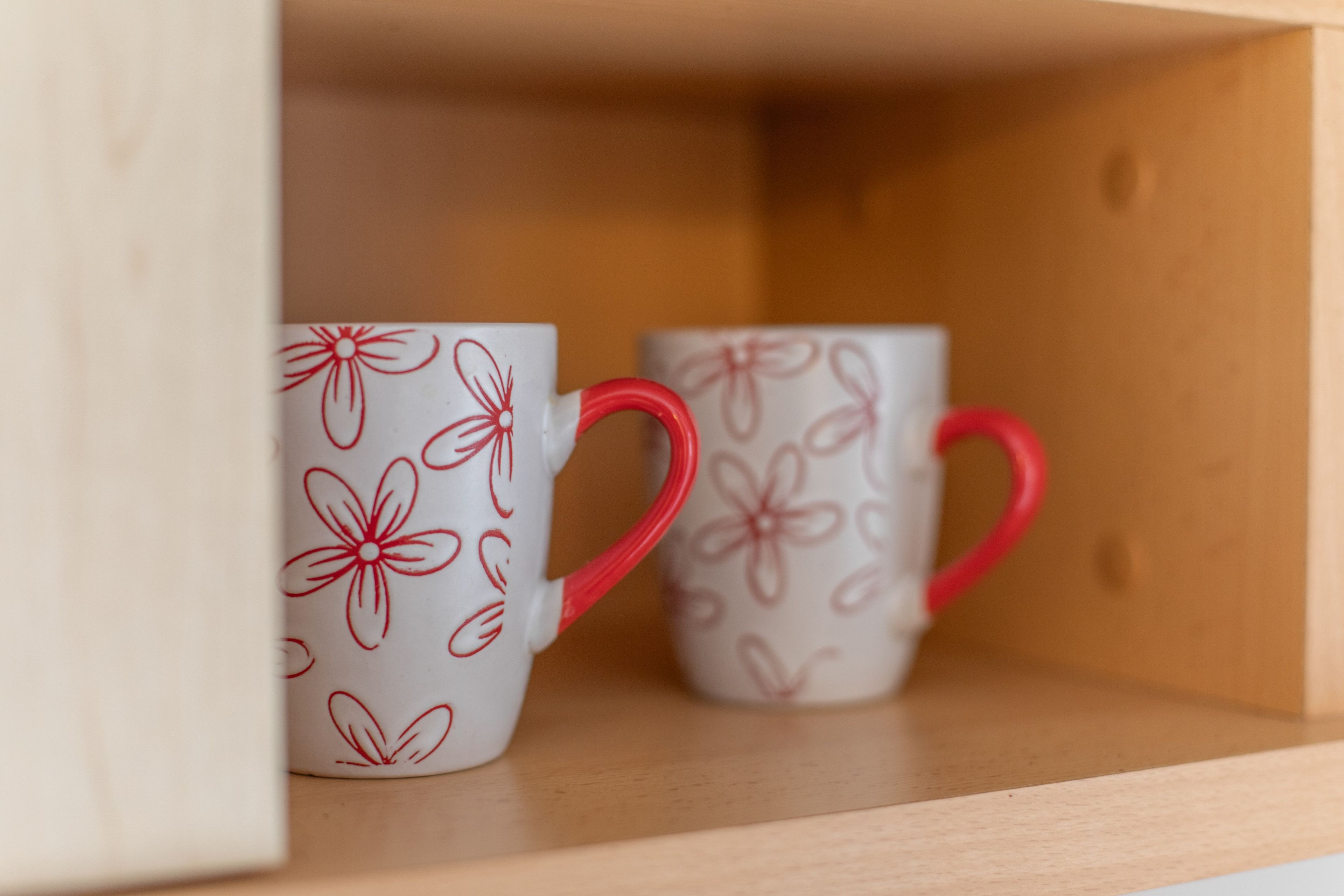 Two mugs with floral patterns and red handles on a shelf.