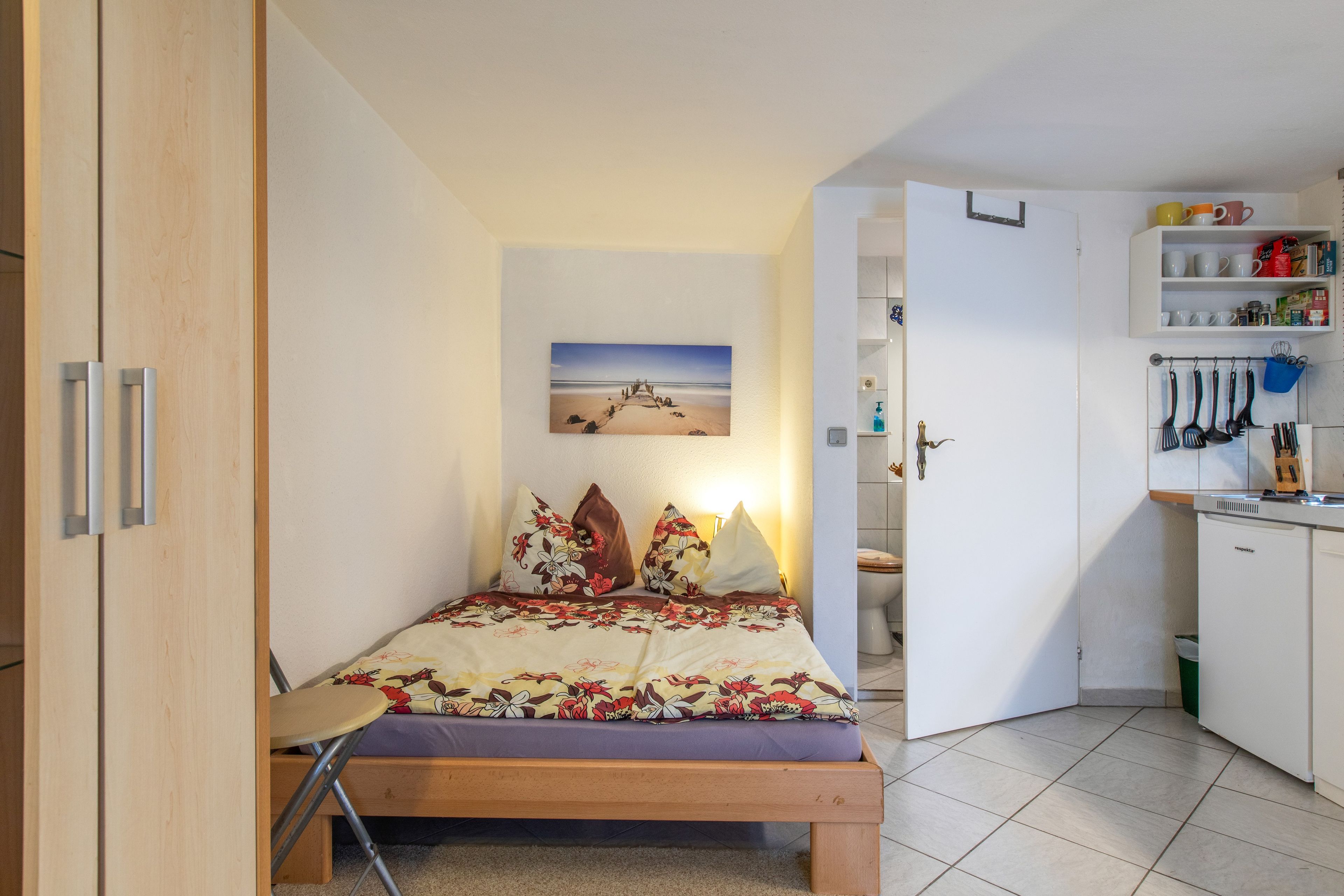 Ein Schlafzimmer mit Bett, Sessel, einem kleinen Tisch, einem Bild an der Wand und einer Küchenzeile mit Geschirr und Spüle.