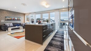 Eine moderne Küche mit einer zentralen Kochinsel, einem Essbereich mit Tisch und Stühlen, einer Couch und einem großen Fenster mit Blick ins Freie.