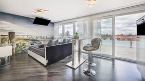 A modern living room with a sofa, a small table, and a bar stool. There is a TV mounted on the wall and a large window offering a view of the city.