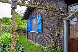 Une maison en bois avec une fenêtre et des volets, entourée de verdure et d’un jardin.