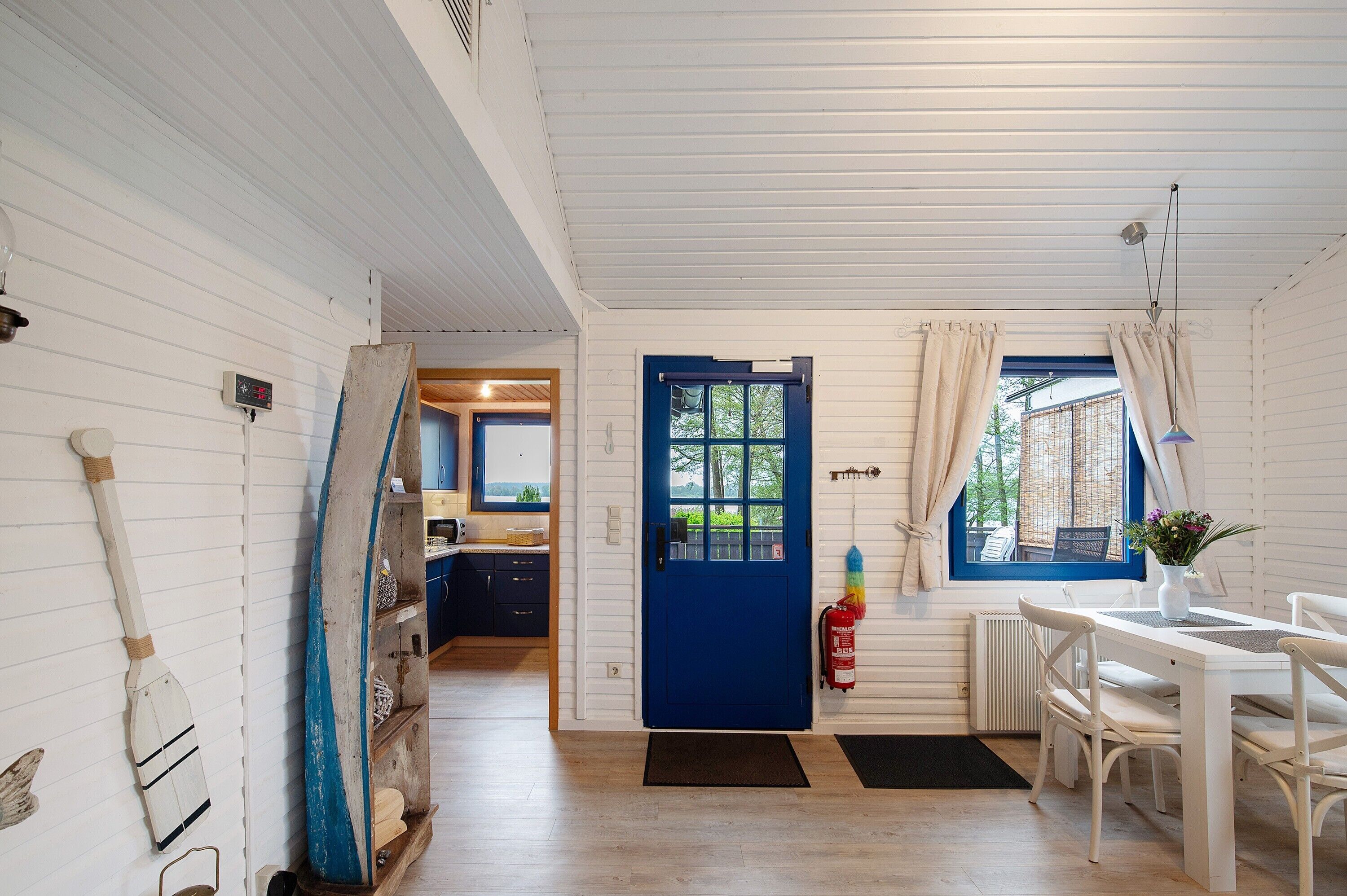 Een kamer met een blauwe deur, witte muren, een eettafel met stoelen, een raam met gordijnen en uitzicht op buiten.