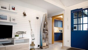Una habitación con televisión, estantes de madera, una puerta azul y una cocina visible a través de un vano abierto.