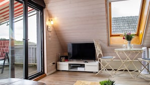 Ein gemütliches Zimmer mit Fernseher, einem kleinen Tisch und einem Sessel. Eine Glastür führt nach draußen.