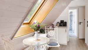 Ein Zimmer mit schrägen Wänden, einem Esstisch mit Glasplatte, einem weißen Stuhl, einem weißen Schrank und Blick durch ein Fenster.