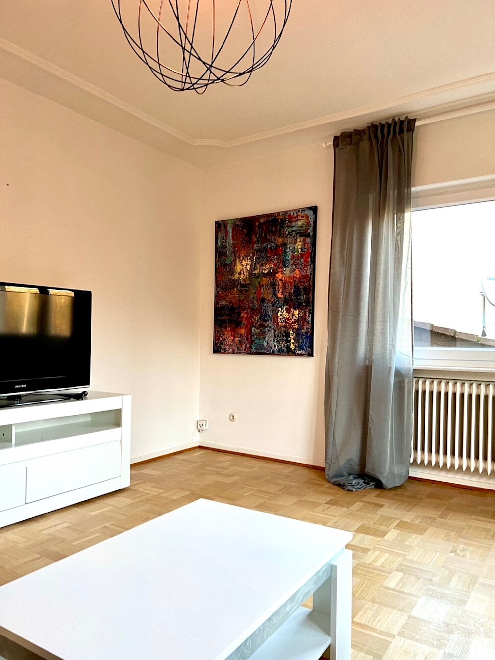 Ein modernes Wohnzimmer mit einem Flachbildfernseher, einem weißen Tisch, einem großen abstrakten Gemälde und einem Fenster mit Vorhängen.