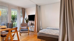 A modern room with a bed, a TV, a dining table with chairs, and a view of greenery outside. - Apartment 'Blackforest Perla' – Cozy Flat with Indoor Pool, Private Terrace, Fast Wi-Fi (Höchenschwand)
