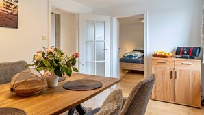 Un comedor con una mesa de madera, sillas, un florero con flores, una cesta y una cama con colchón y almohadas al fondo.