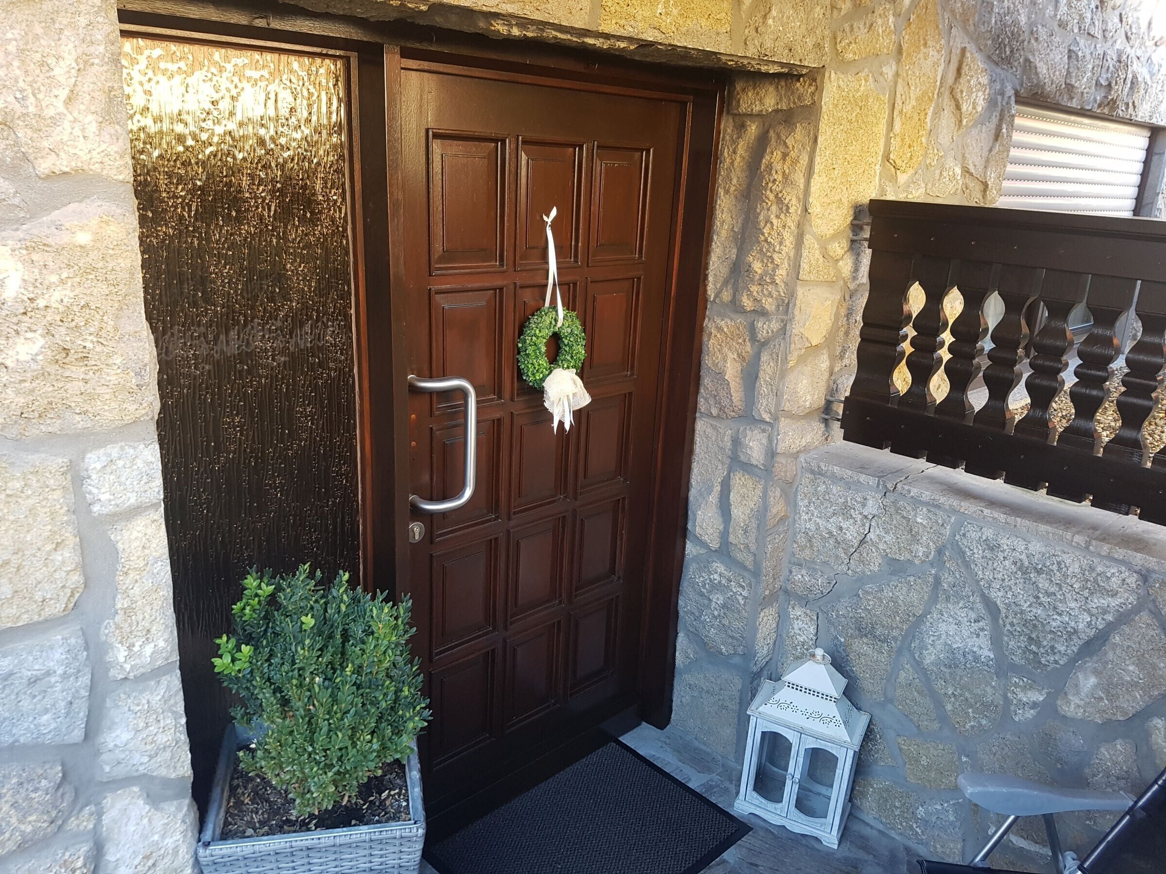 A stone wall, a wooden door with a metal handle, a wreath, a potted plant, and a white lantern.