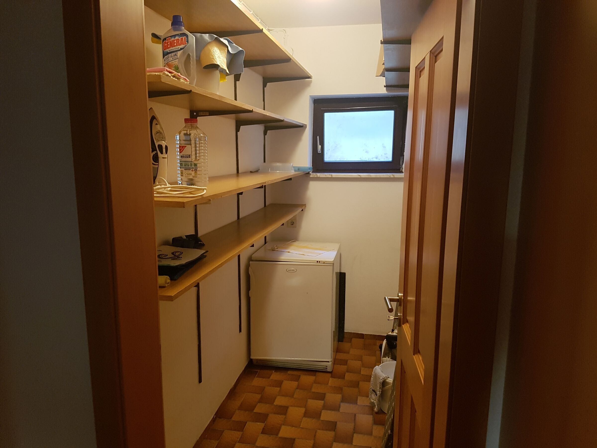 A compact kitchen with a refrigerator, shelves with various items, a window, and a door.