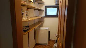 A compact kitchen with a refrigerator, shelves with various items, a window, and a door.