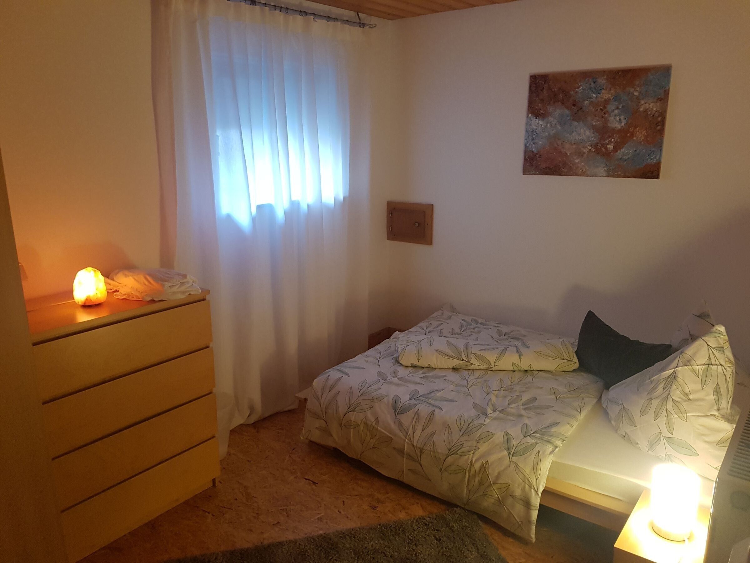 A bedroom with a bed, a nightstand, a dresser, a window with curtains, and a painting on the wall.