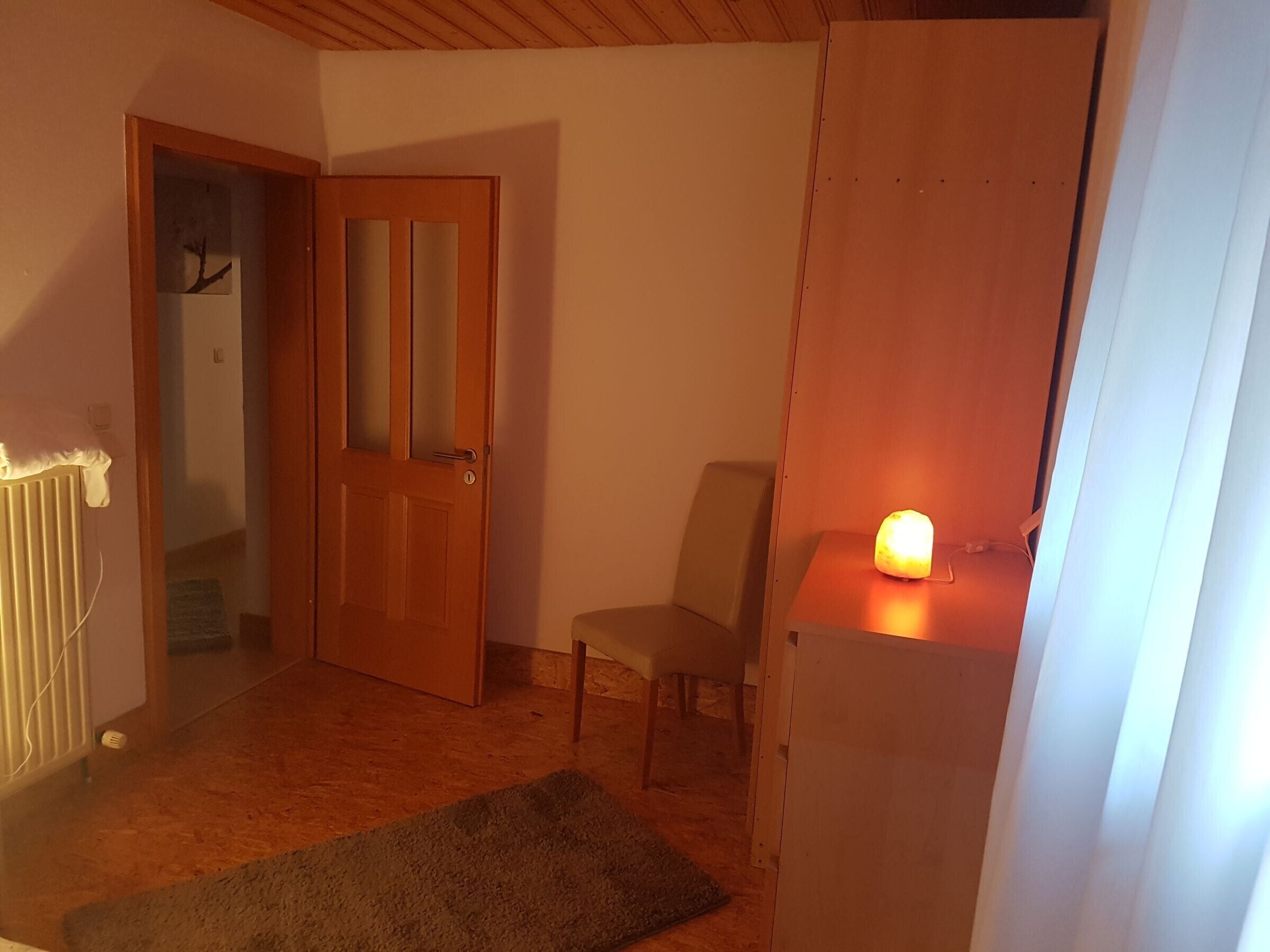 Huoneessa on puinen ovi, patteri, tuoli ja pöydällä oleva, sytytetty lamppu.