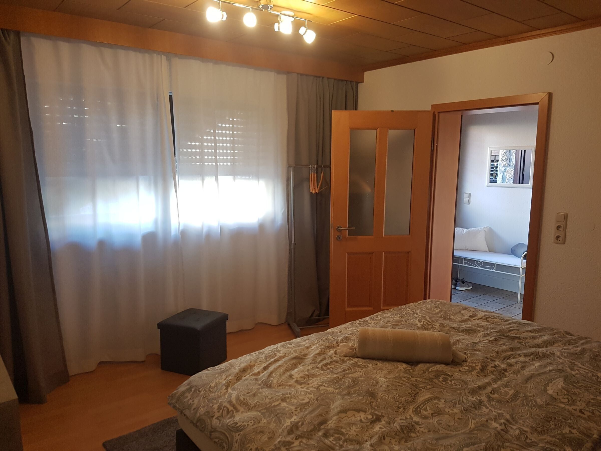 Ein Schlafzimmer mit Bett, Vorhängen, einem kleinen Hocker und einer Tür zu einem anderen Raum.