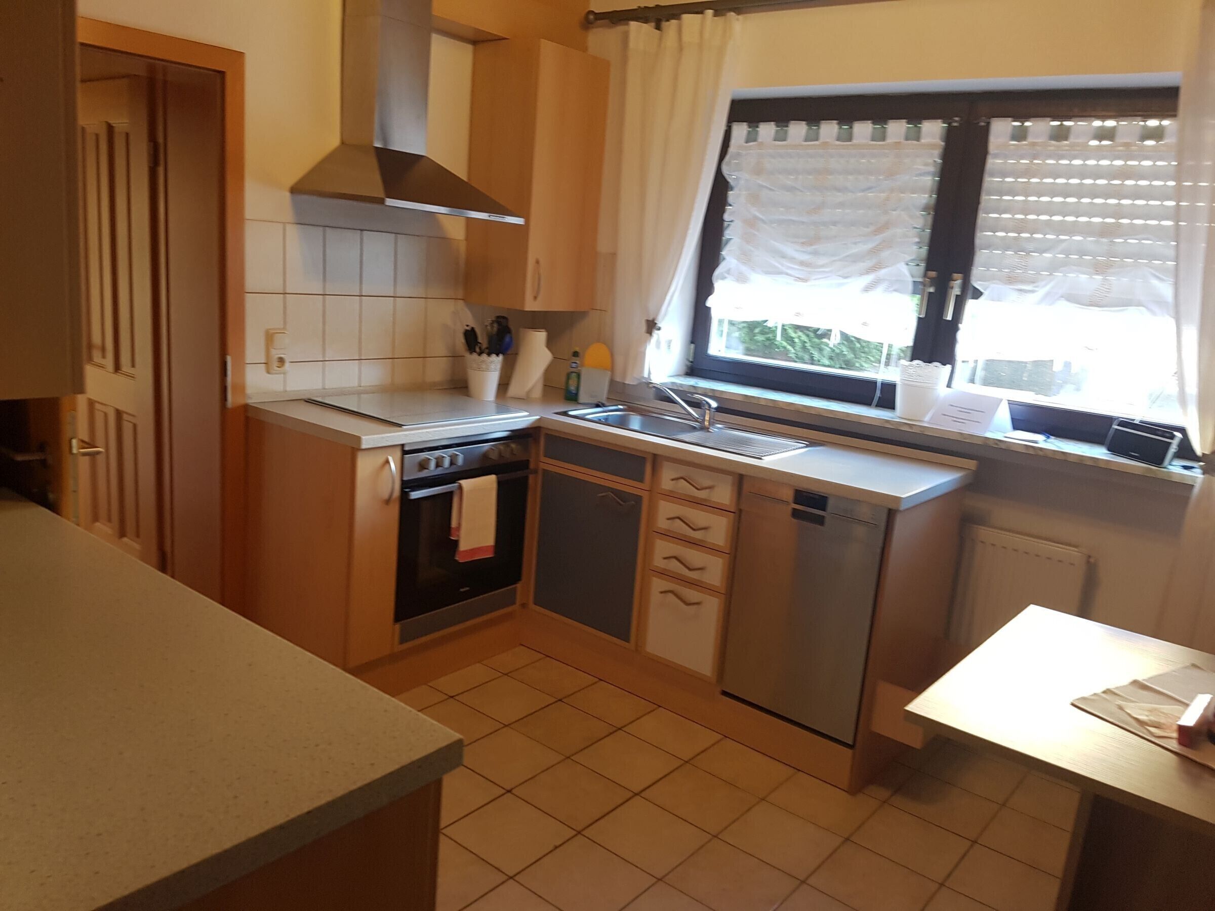 A compact kitchen with a stove, oven, dishwasher, and a window with curtains.