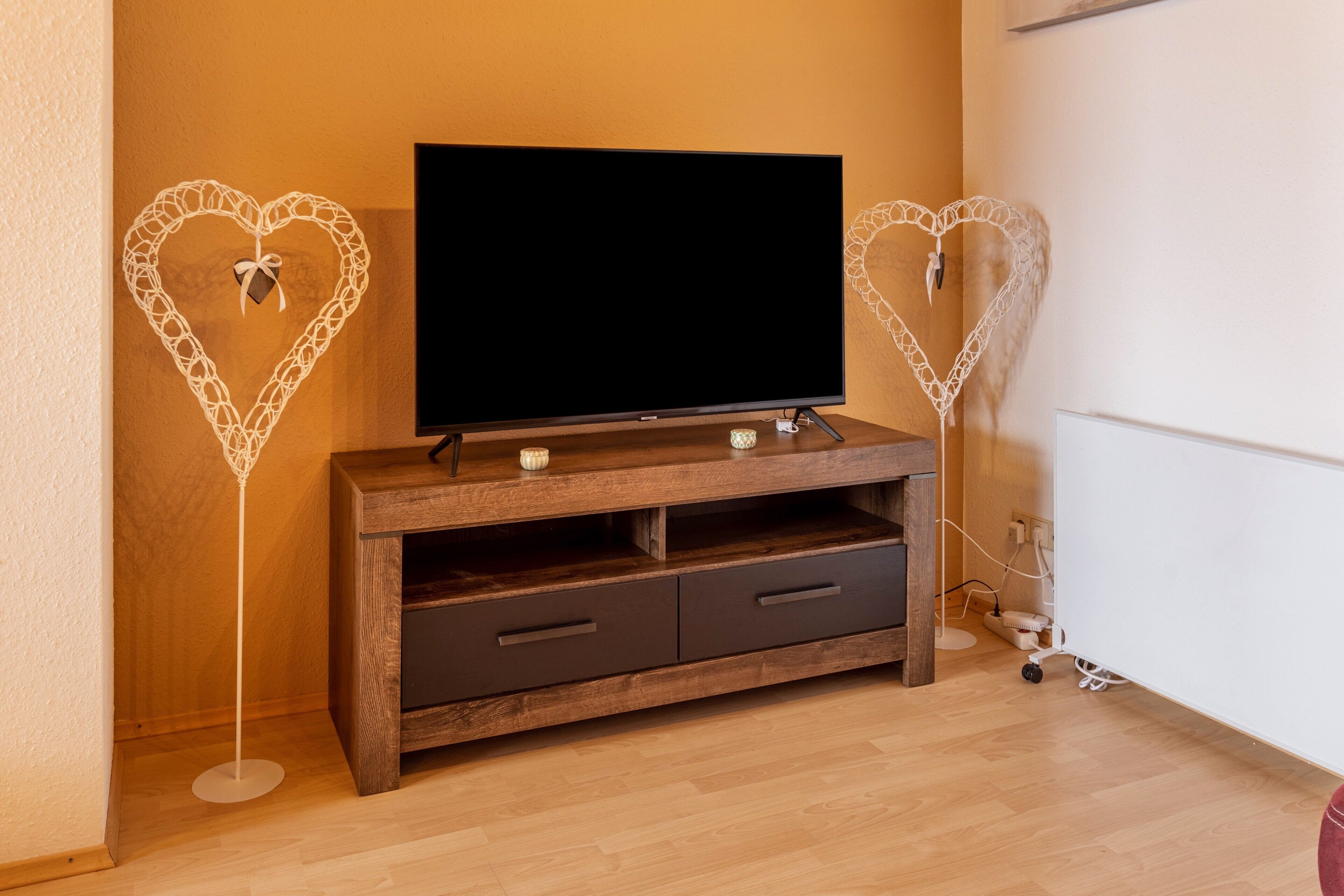 Una sala de estar moderna con un televisor de pantalla plana sobre un mueble de madera, dos adornos en forma de corazón y un radiador blanco.