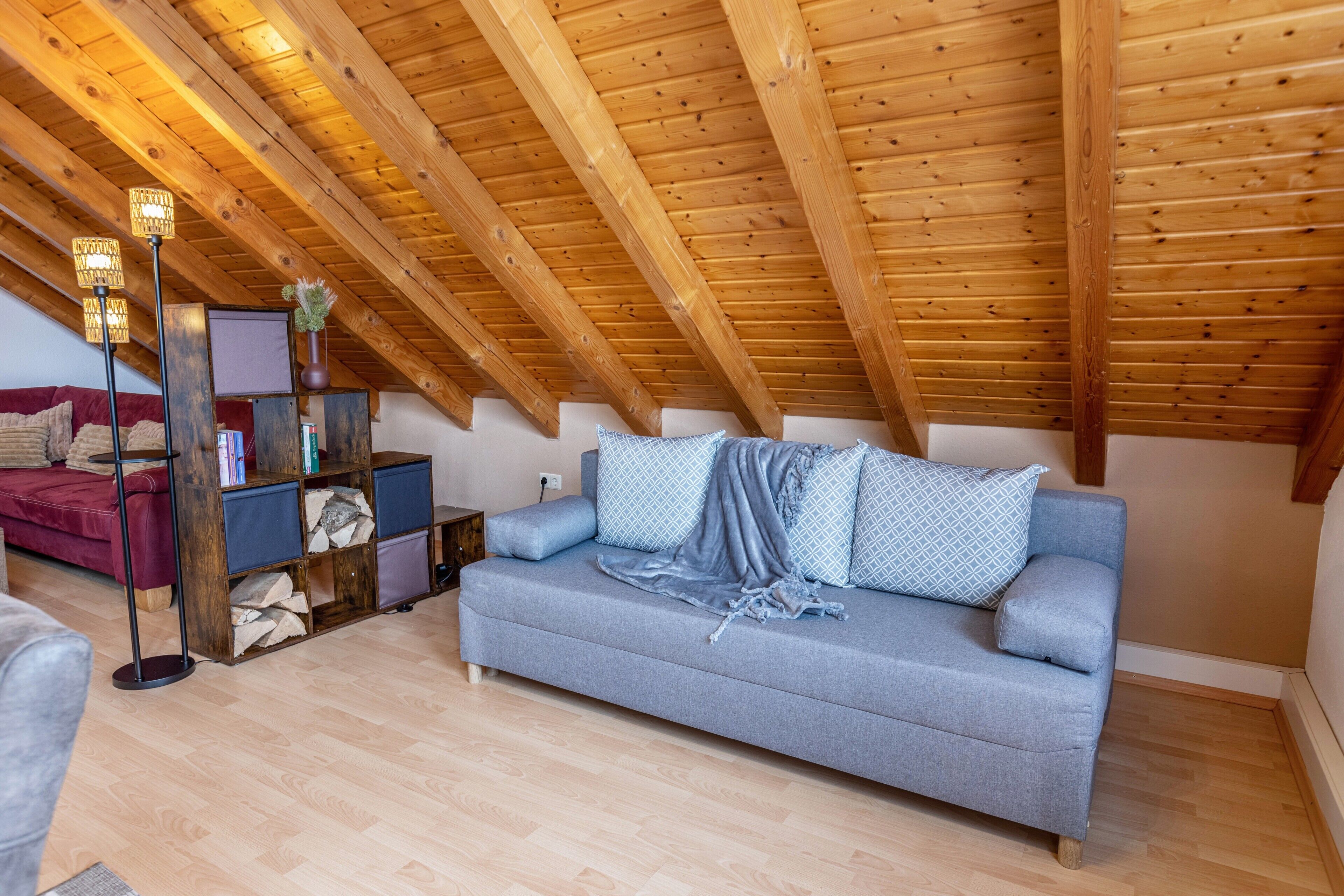 Un espacio tipo loft con techo inclinado, un sofá gris, una lámpara de pie y una estantería.