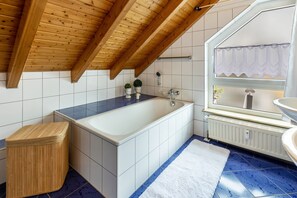 Une salle de bain avec une baignoire, un tabouret en bois, des carreaux blancs, une fenêtre avec vue et un radiateur.