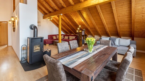 A spacious loft with a high wooden ceiling, a central dining table, a sofa set, and a wood-burning stove.