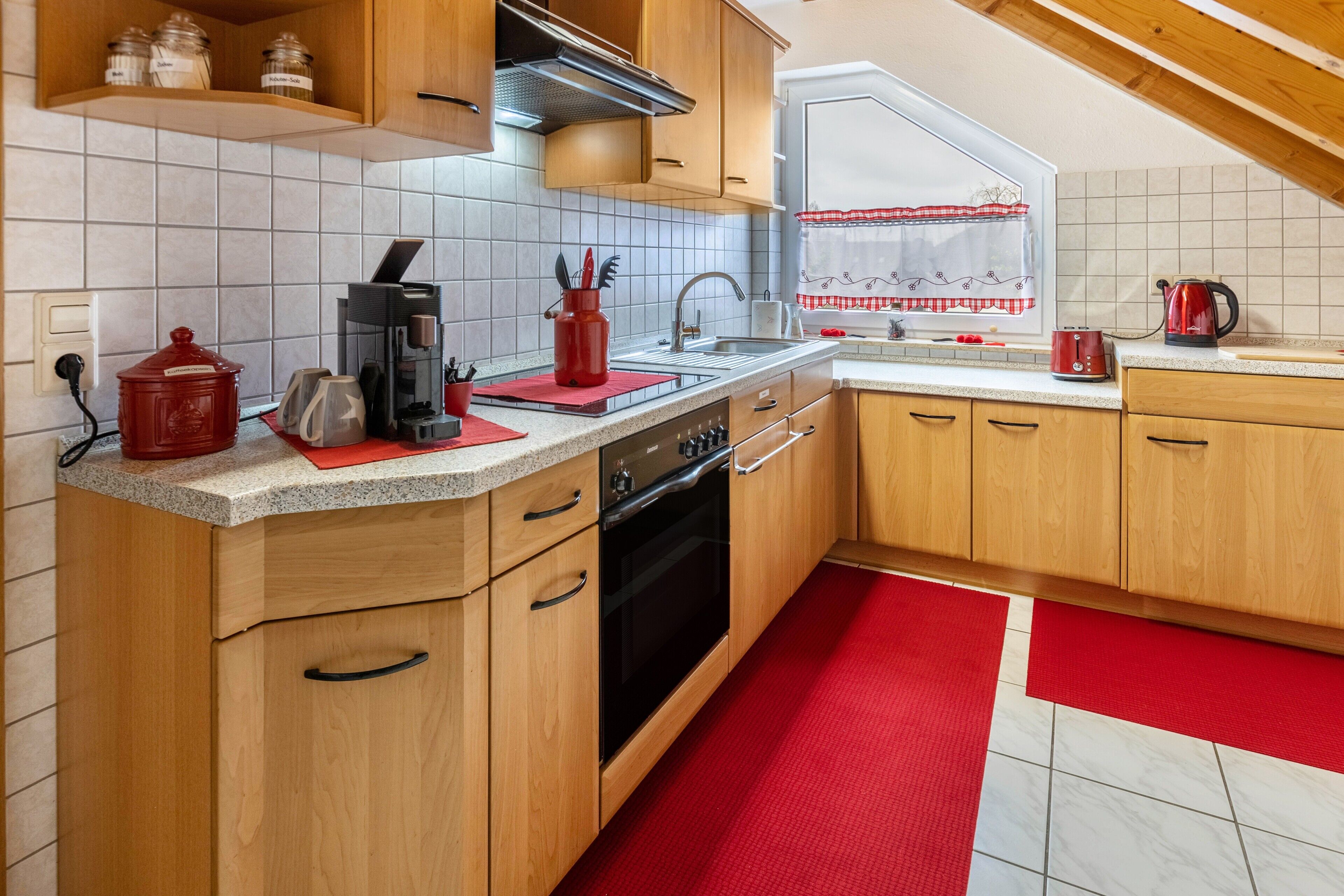 Une cuisine avec des meubles de rangement en bois, une cuisinière, un évier et une fenêtre ornée d’un rideau rouge.
