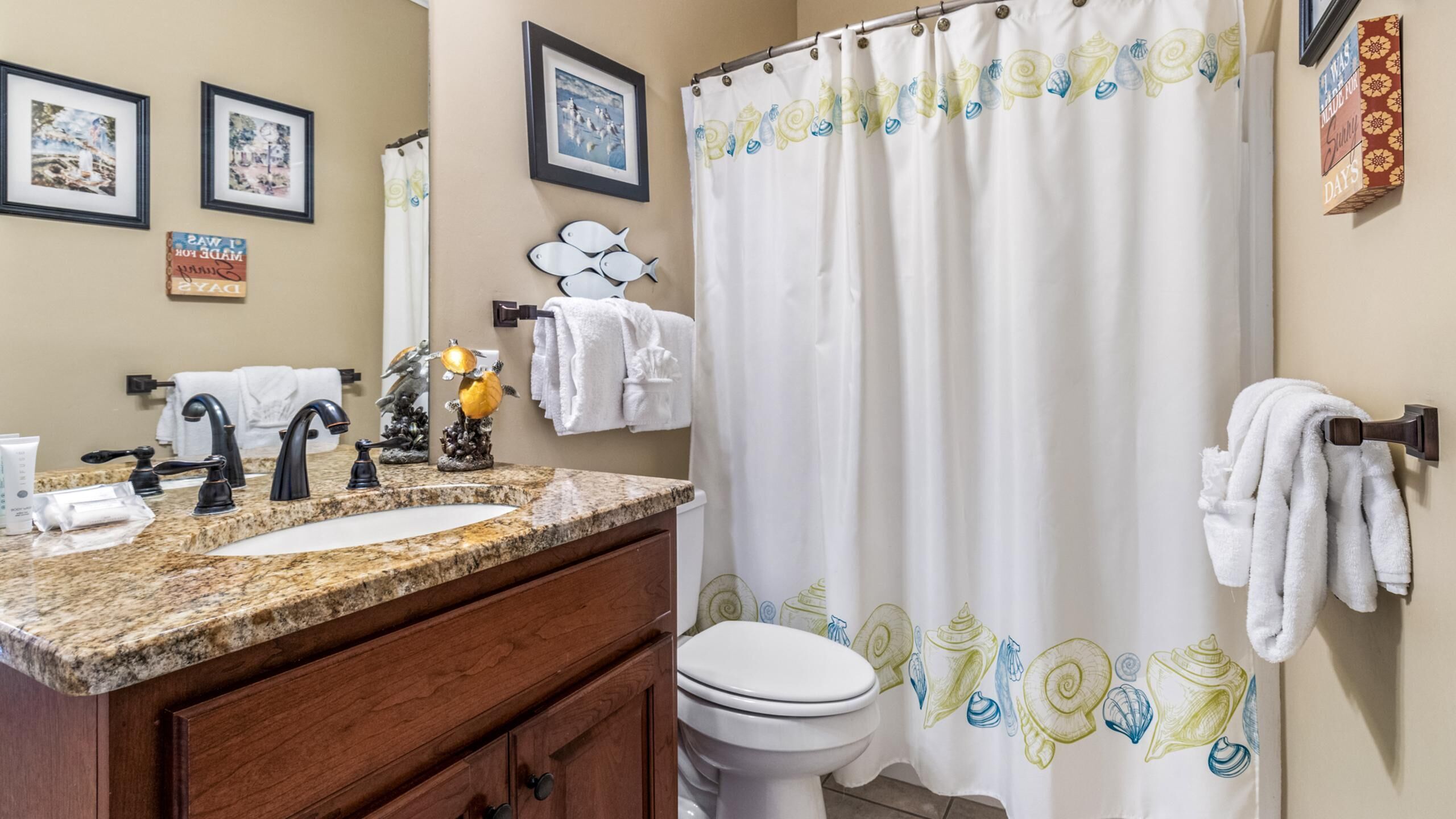 A bathroom with a granite countertop sink, two black faucets, a white toilet, and a shower curtain with a shell pattern.