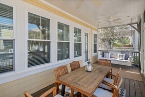 A covered patio with a wooden table and chairs, large windows with blinds, and a view of the outdoors.
