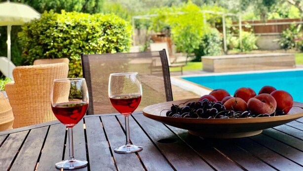 Une table en bois, avec deux verres à vin et un saladier de fruits, placée au bord d’une piscine.
