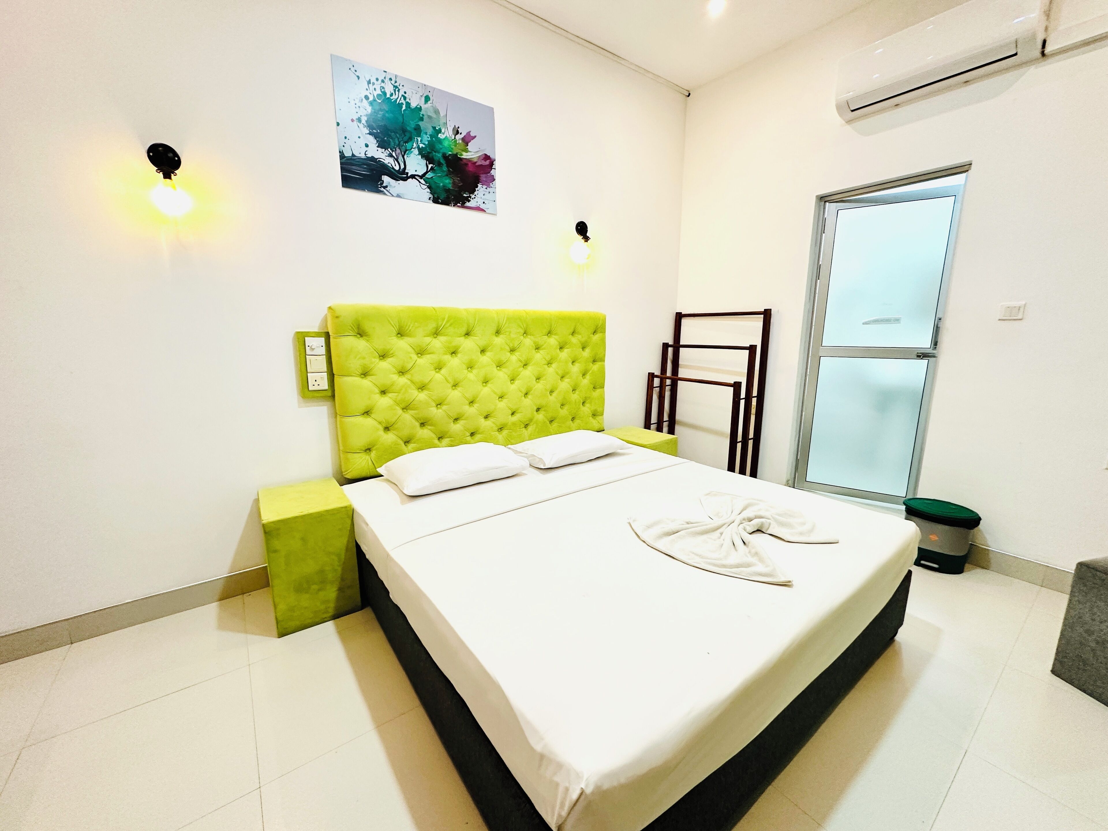 Una cama bien hecha con cabecera verde, una mesita de noche, una lámpara de pared y una ventana con vistas.