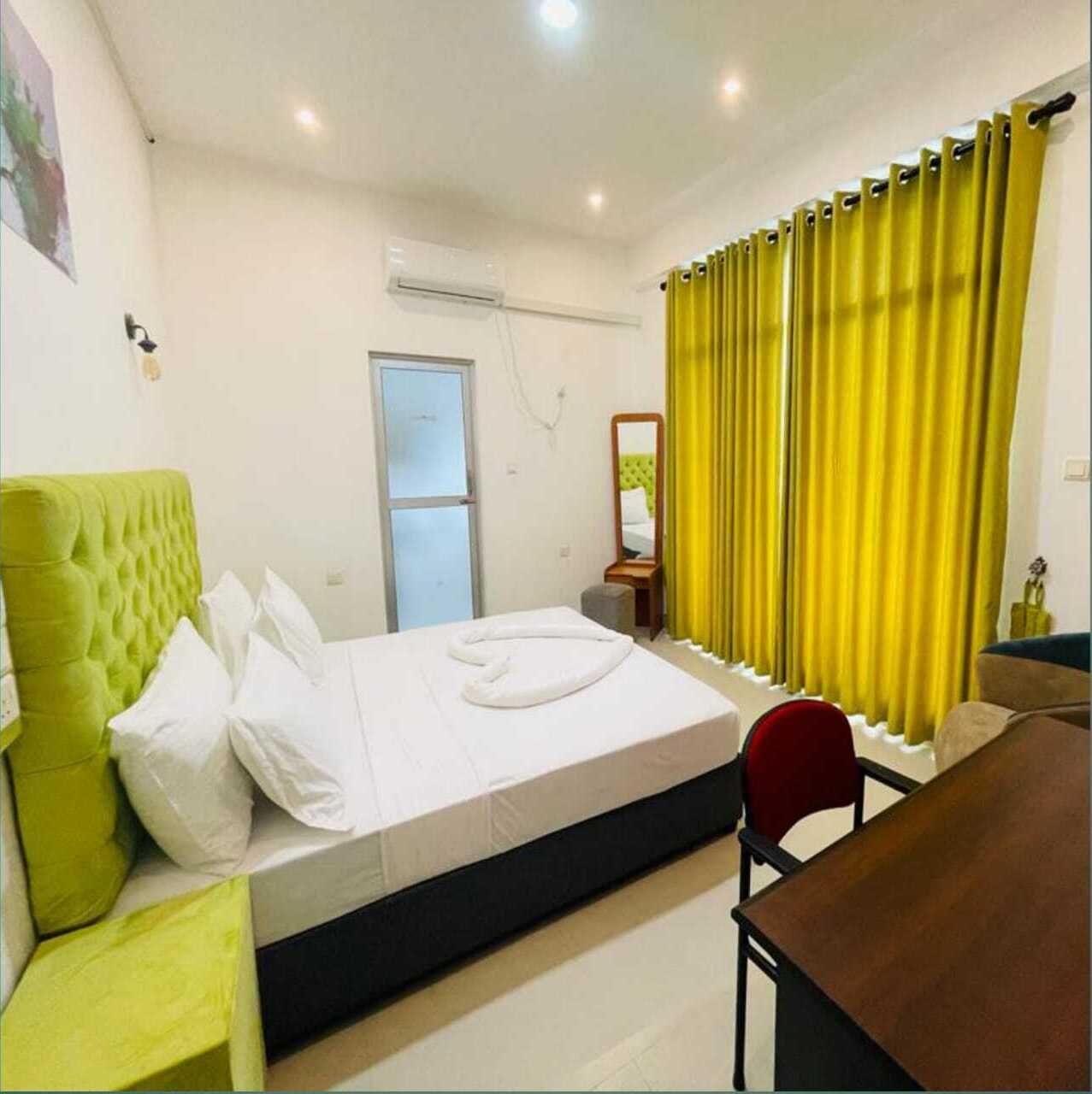 Une chambre d’hôtel avec un lit, une tête de lit verte, une table de chevet en bois, un miroir et des rideaux jaunes.
