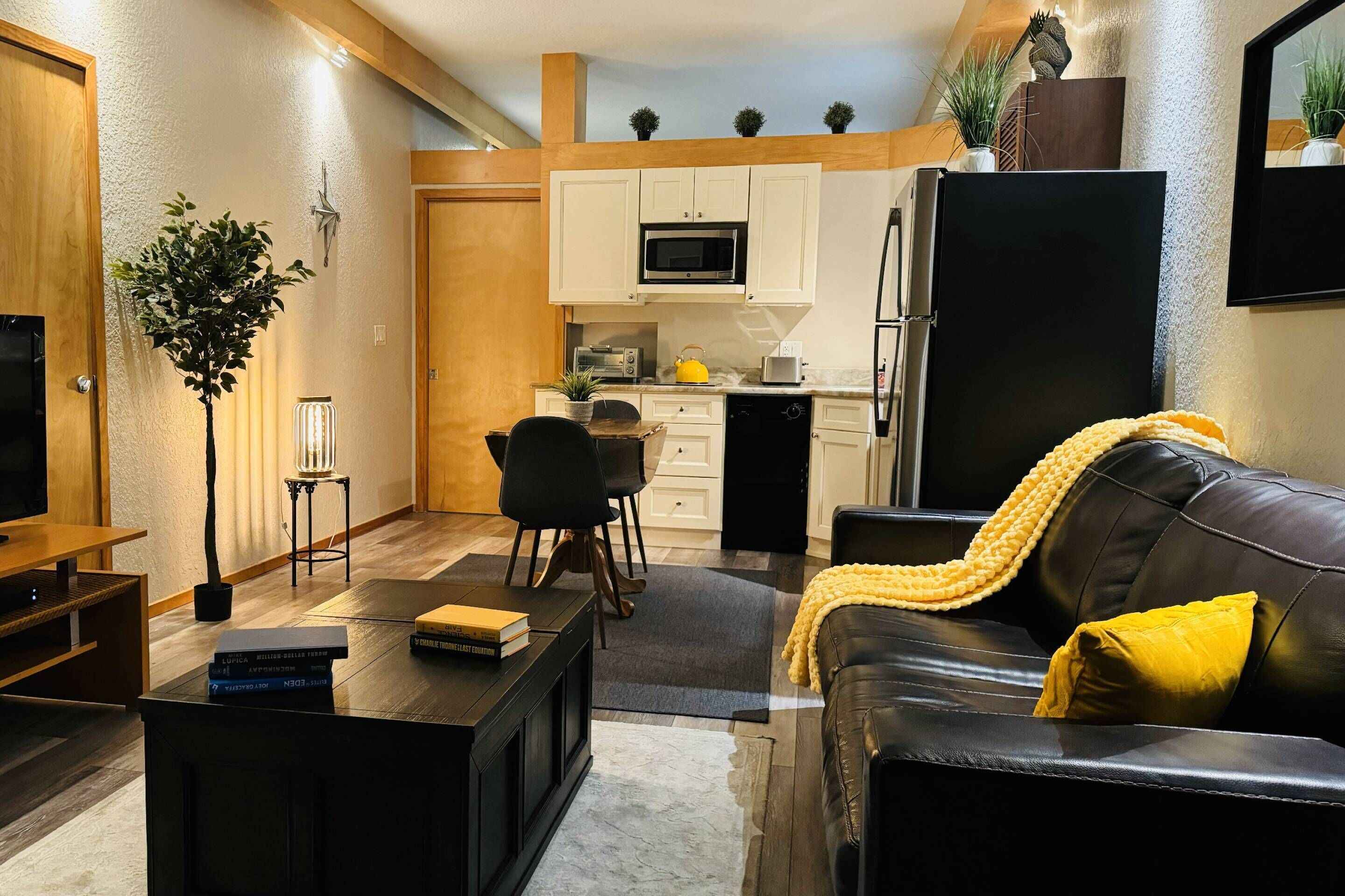 A compact living space with a kitchenette, a black leather sofa with a yellow throw, a wooden coffee table with books, and a TV on a wooden stand.
